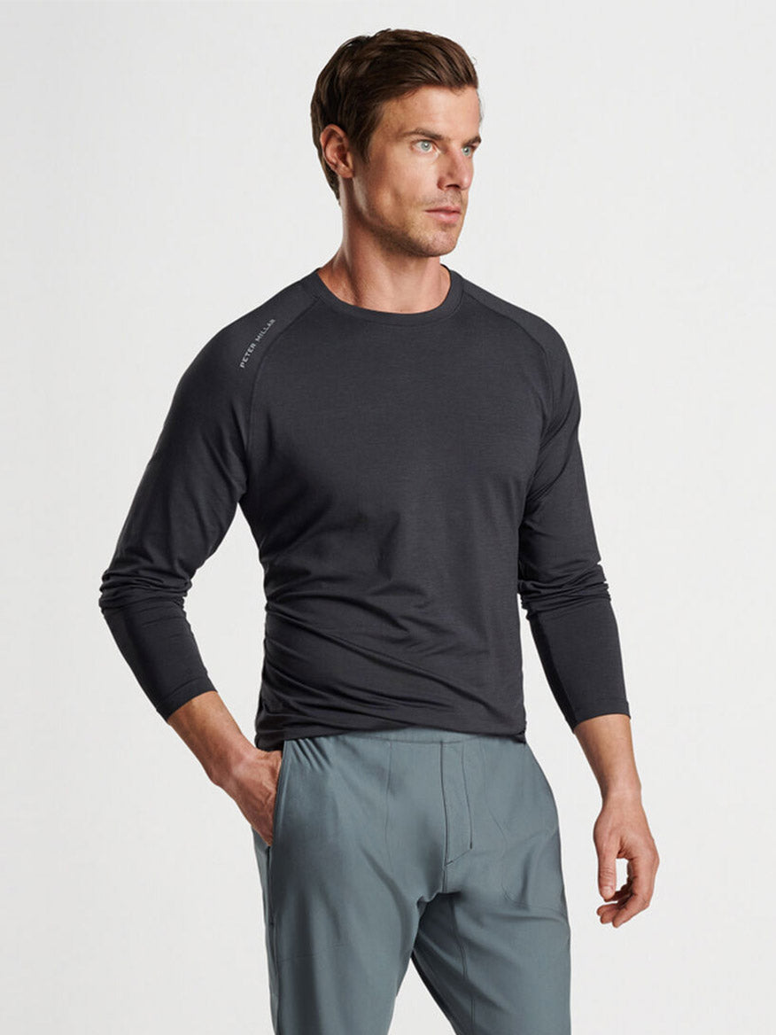 A man wearing a Peter Millar Aurora Performance Long-Sleeve T-shirt in Black with UPF 50+ sun protection and light-colored pants standing with one hand on his hip.