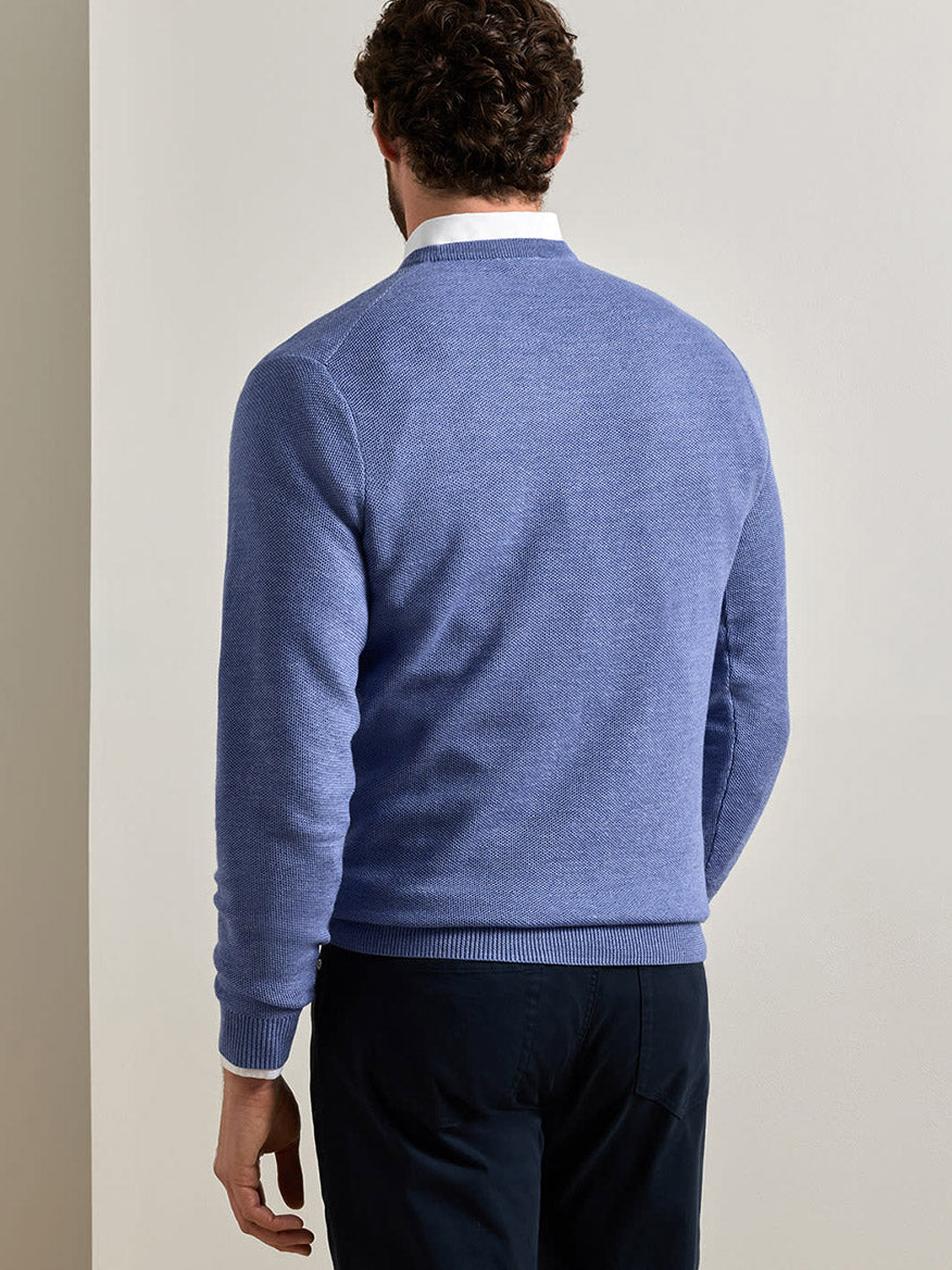 A man stands facing away from the camera, wearing the Peter Millar Autumn Crest Honeycomb Crewneck Sweater in Blue Freesia—a classic fit, light blue sweater with a subtle honeycomb texture—layered over a white collared shirt and paired with dark trousers against a plain, light-colored background.