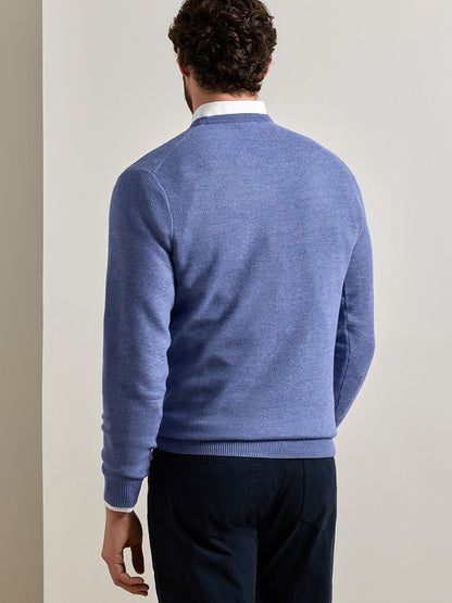 A man stands facing away from the camera, wearing the Peter Millar Autumn Crest Honeycomb Crewneck Sweater in Blue Freesia—a classic fit, light blue sweater with a subtle honeycomb texture—layered over a white collared shirt and paired with dark trousers against a plain, light-colored background.