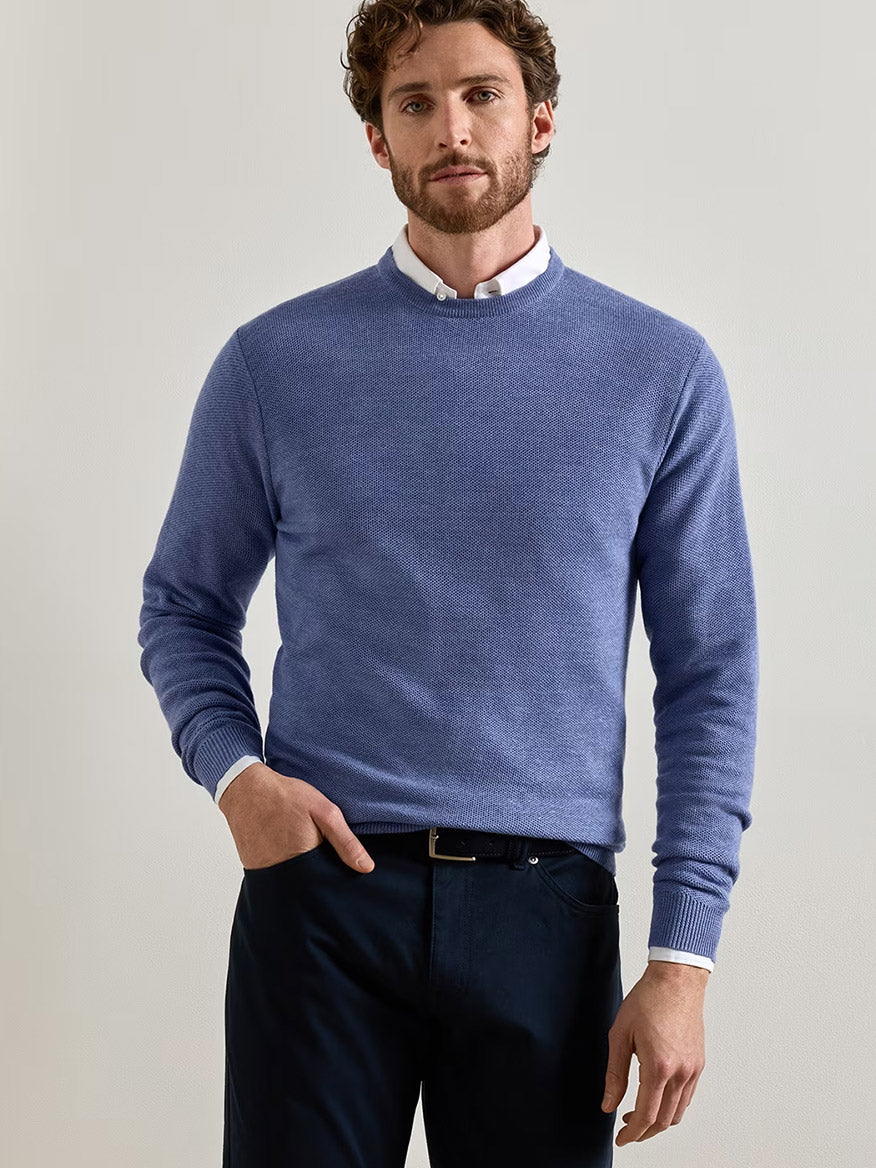 A man wearing the Peter Millar Autumn Crest Honeycomb Crewneck Sweater in Blue Freesia over a white collared shirt and dark pants, standing against a plain light background.