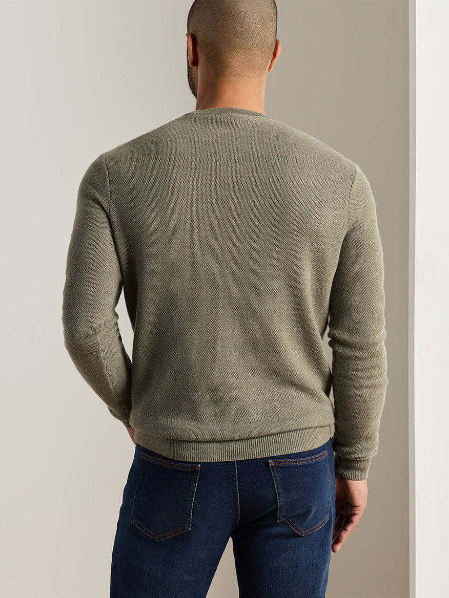 A person stands facing away from the camera, wearing the Peter Millar Autumn Crest Honeycomb Crewneck Sweater in Fatigue—a light olive green sweater in a classic fit—paired with blue jeans, in a neutral indoor setting.