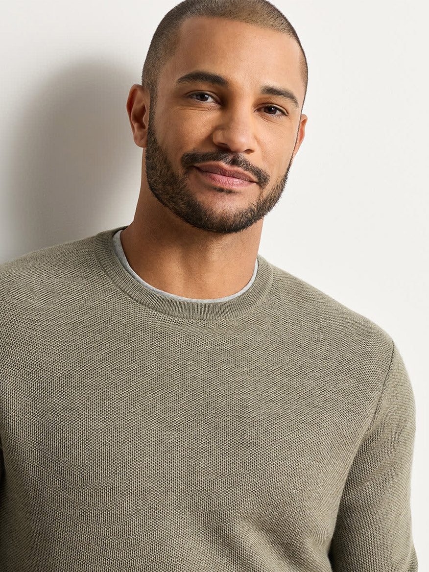 A man with short hair and a trimmed beard wears the Peter Millar Autumn Crest Honeycomb Crewneck Sweater in Fatigue, featuring a classic fit and subtle honeycomb texture, standing against a plain light background and looking at the camera with a slight smile.