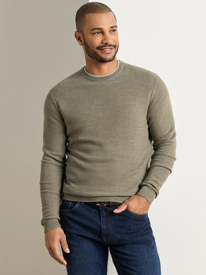 A man stands against a plain background wearing the Peter Millar Autumn Crest Honeycomb Crewneck Sweater in Fatigue—a classic fit crewneck sweater with a subtle honeycomb texture—paired with blue jeans, looking off to the side with one hand in his pocket.