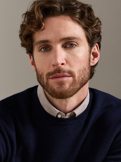 A man with curly brown hair, a beard, and light eyes models the Peter Millar Autumn Crest Honeycomb Crewneck Sweater in Navy—a classic fit crewneck featuring a subtle honeycomb texture—layered over a button-up shirt, as he looks directly at the camera against a plain background.