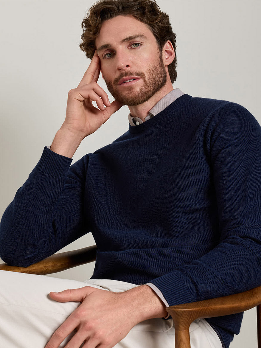 A man with short curly hair and a beard sits in a wooden chair, wearing the Peter Millar Autumn Crest Honeycomb Crewneck Sweater in Navy over a light button-up shirt and white pants, with one hand resting on his face.
