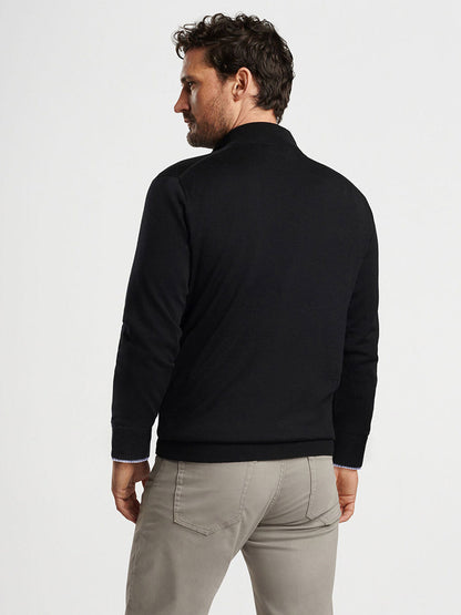 A man with curly hair is standing and facing away, wearing a sleek black Peter Millar Autumn Crest Quarter-Zip sweater and beige pants against a plain white background.