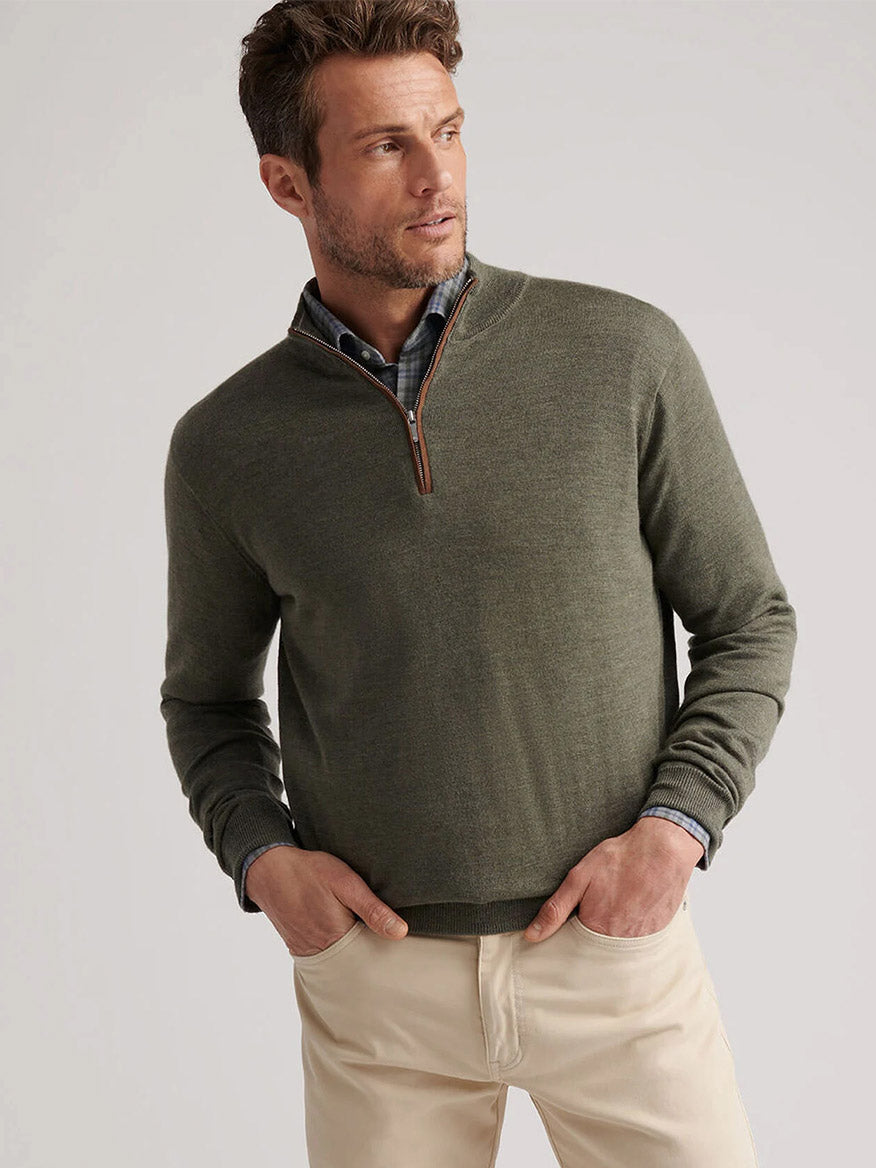 A man wears the Peter Millar Autumn Crest Suede Trim Quarter-Zip in Basil over a button-up shirt and beige pants, standing with his hands in his pockets against a plain background.