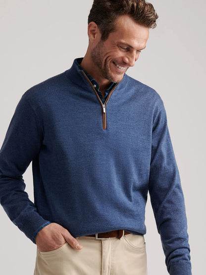 A man wears the Peter Millar Autumn Crest Suede Trim Quarter-Zip in Blue Haze over a collared shirt and beige pants, standing with one hand in his pocket as he looks down and smiles.
