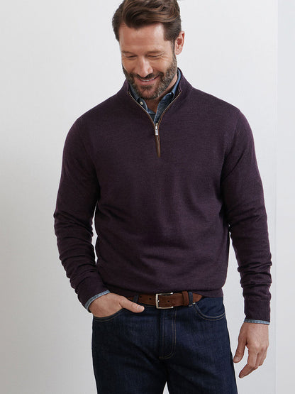 A man wears the Peter Millar Autumn Crest Suede Trim Quarter-Zip in Midnight Grape over a button-down shirt and jeans, standing against a plain light-colored wall while looking down and smiling.