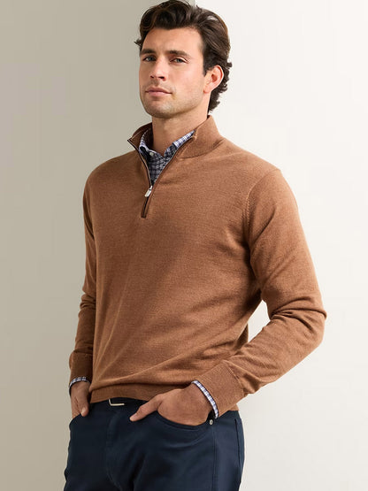 A man stands against a plain off-white background with one hand in his pocket, wearing the Peter Millar Autumn Crest Suede Trim Quarter-Zip in Dark Amber layered over a checked shirt and paired with navy pants.