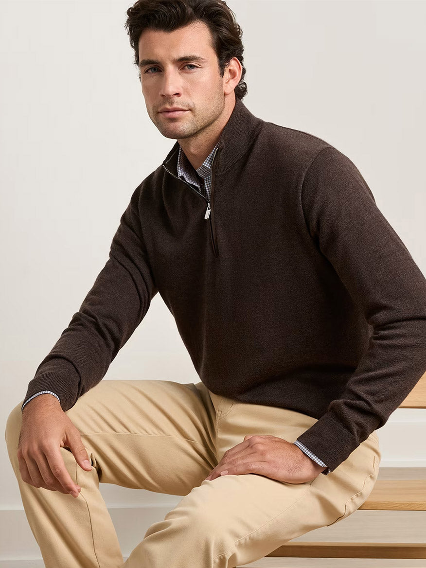 A man seated on a wooden bench wears the Peter Millar Autumn Crest Suede Trim Quarter-Zip in French Press over a collared shirt and beige pants against a plain background.