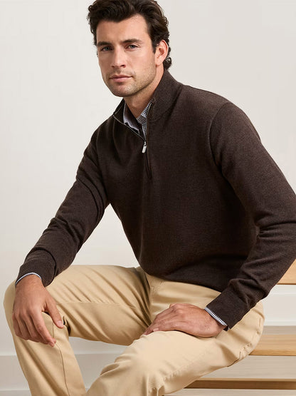 A man seated on a wooden bench wears the Peter Millar Autumn Crest Suede Trim Quarter-Zip in French Press over a collared shirt and beige pants against a plain background.
