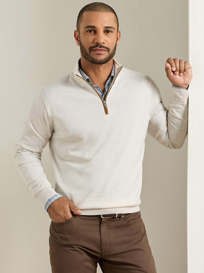 A man with short hair and a beard is wearing a Peter Millar Autumn Crest Suede Trim Quarter-Zip in Ivory over a blue shirt, standing indoors with one hand in his pocket and the other resting against a wall.