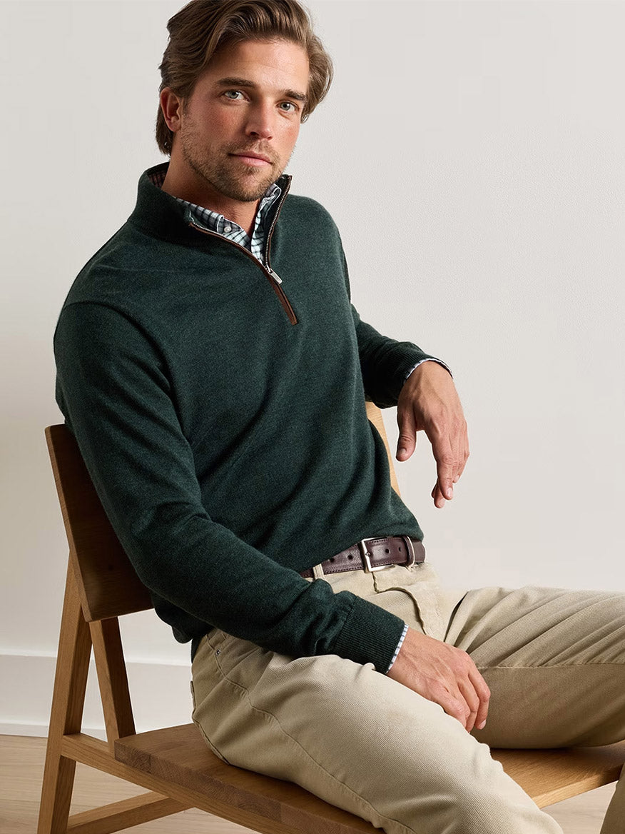 A man sits on a wooden chair wearing the Peter Millar Autumn Crest Suede Trim Quarter-Zip in Pine Brook over a plaid shirt and beige pants, looking at the camera with a neutral expression.