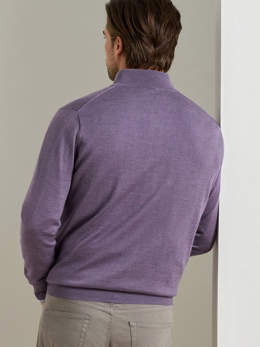 A man with brown hair, wearing a Peter Millar Autumn Crest Suede Trim Quarter-Zip in Purple Fig and gray pants, stands with his back to the camera and leans against a white wall.