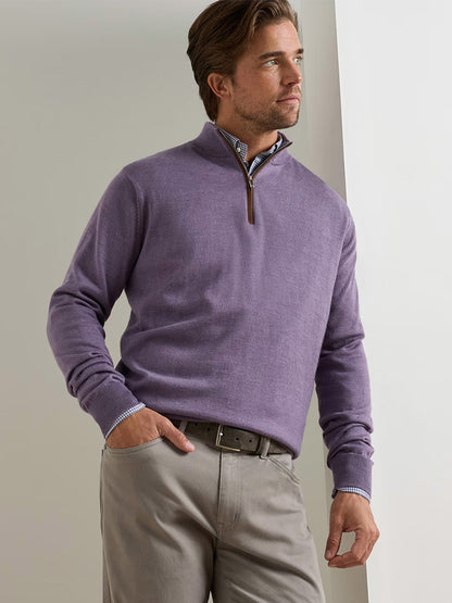 A man stands near a white wall wearing the Peter Millar Autumn Crest Suede Trim Quarter-Zip in Purple Fig over a checkered shirt, paired with beige pants and a brown belt. This lightweight sweater combines comfort and style.