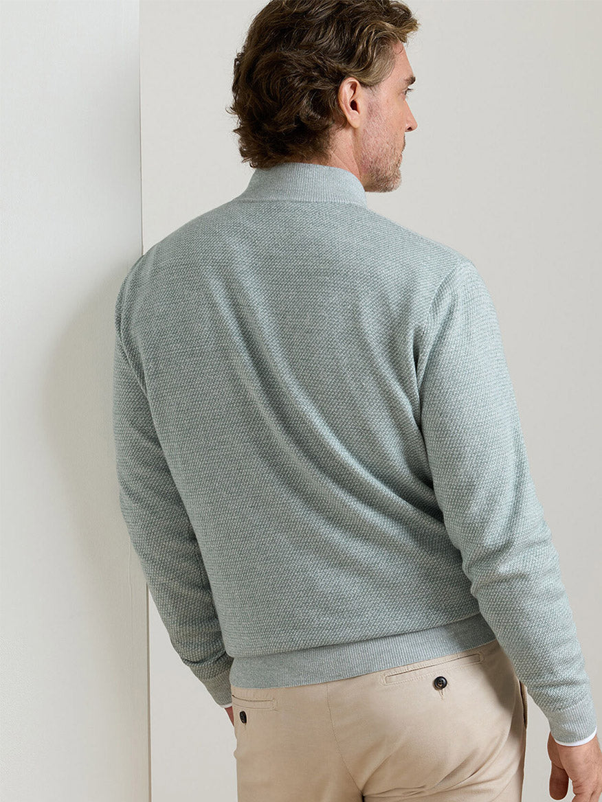 A man stands facing away in a minimal indoor setting, wearing the Peter Millar Barlow Quarter-Zip Sweater in Willow and beige pants.