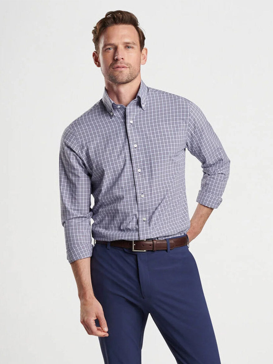 A man with short hair wears a Peter Millar Berkshire Performance Poplin Sport Shirt in navy with blue and white checks, crafted from soft performance fabric. His dark blue pants complement the look, and his sleeves are rolled up. With his left hand in his pocket, he exudes casual confidence while enjoying UPF 50+ sun protection.