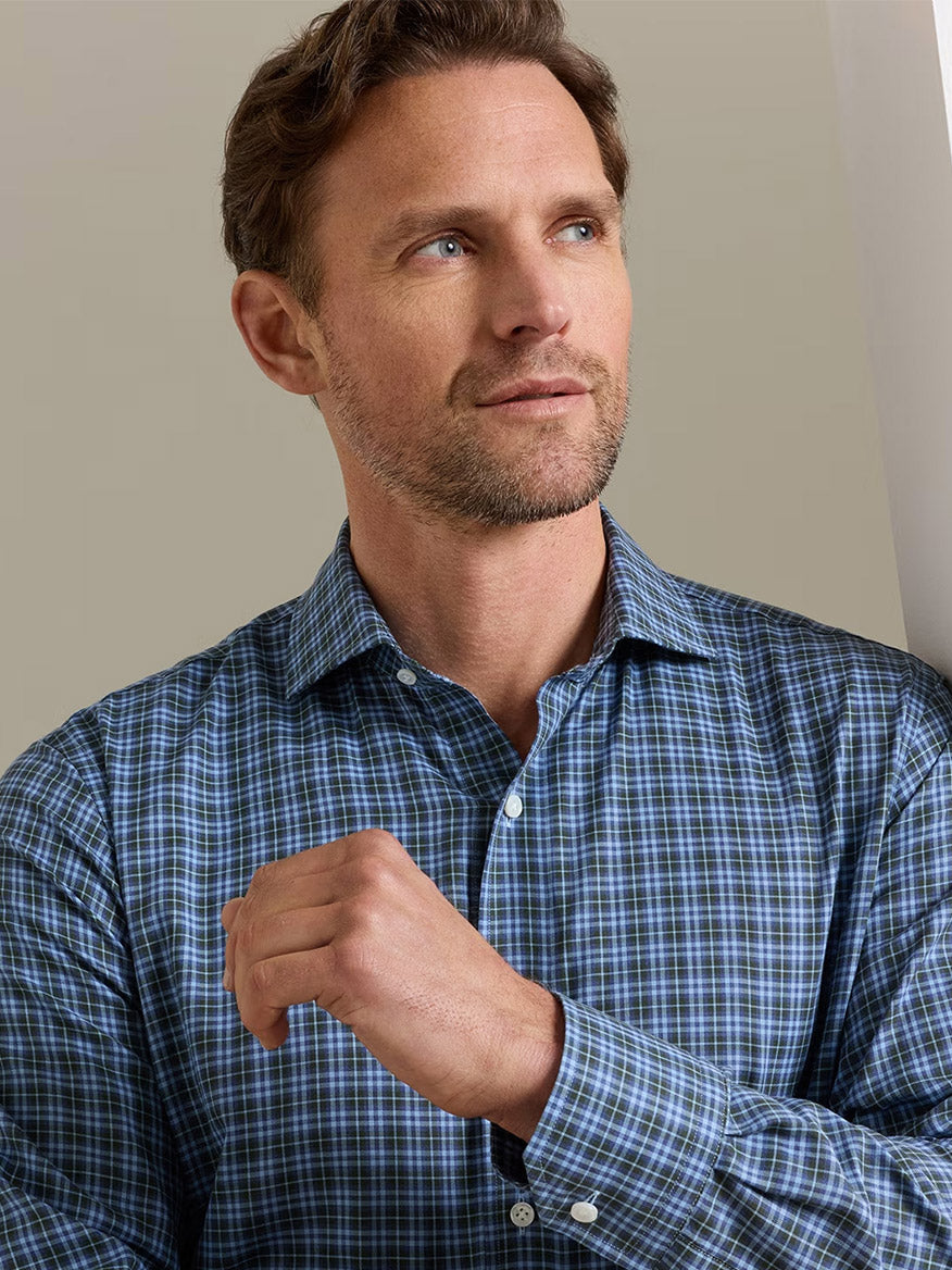 A man with short brown hair and a beard wears the Peter Millar Bernard Cotton Sport Shirt in Windy Blue—a tailored-fit, blue plaid shirt made from cotton poplin—standing indoors and looking slightly to the side with his hand raised near his chest.