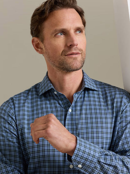 A man with short brown hair and a beard wears the Peter Millar Bernard Cotton Sport Shirt in Windy Blue—a tailored-fit, blue plaid shirt made from cotton poplin—standing indoors and looking slightly to the side with his hand raised near his chest.