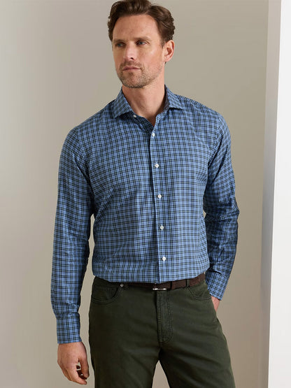 A man wearing the Peter Millar Bernard Cotton Sport Shirt in Windy Blue and olive green pants stands against a plain beige wall.