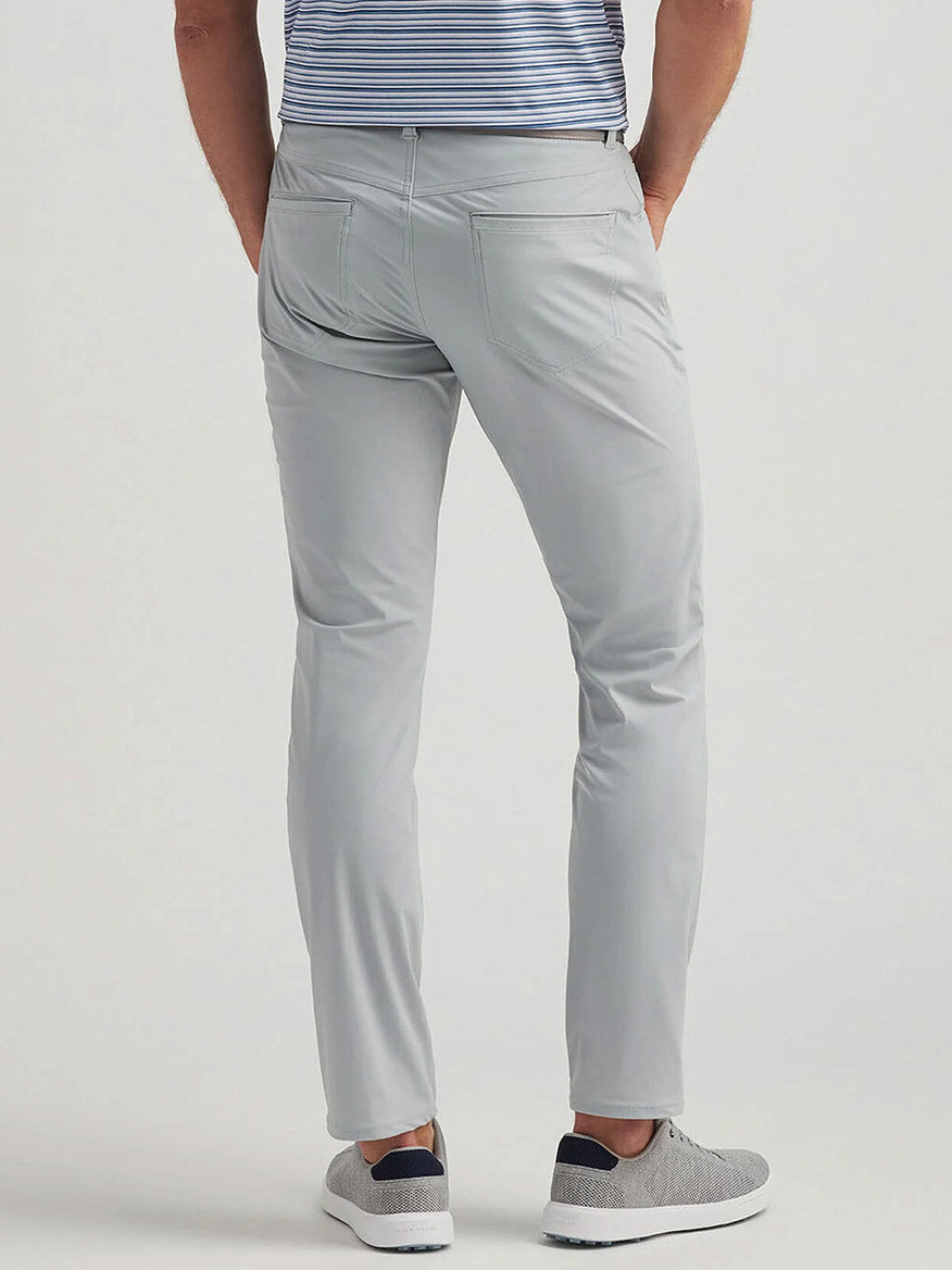 A man stands with his back to the camera, wearing Peter Millar Bingham Performance Five-Pocket Pant in British Grey, a blue and white striped shirt, and gray slip-on shoes.