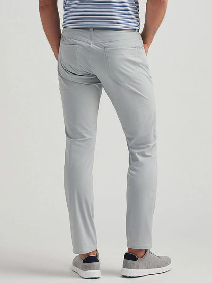 A man stands with his back to the camera, wearing Peter Millar Bingham Performance Five-Pocket Pant in British Grey, a blue and white striped shirt, and gray slip-on shoes.