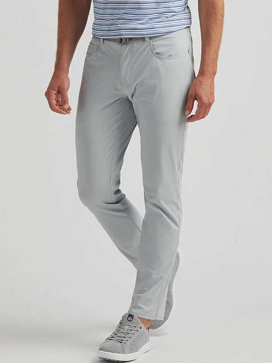 A person wearing Peter Millar Bingham Performance Five-Pocket Pant in British Grey, a blue and white striped shirt, and grey sneakers stands against a plain background.