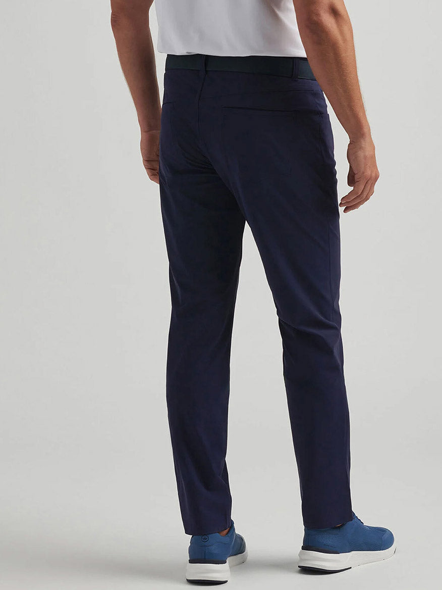 A man in Peter Millar Bingham Performance Five-Pocket Pant in Navy, a white shirt, and blue sneakers stands facing away from the camera against a plain background.