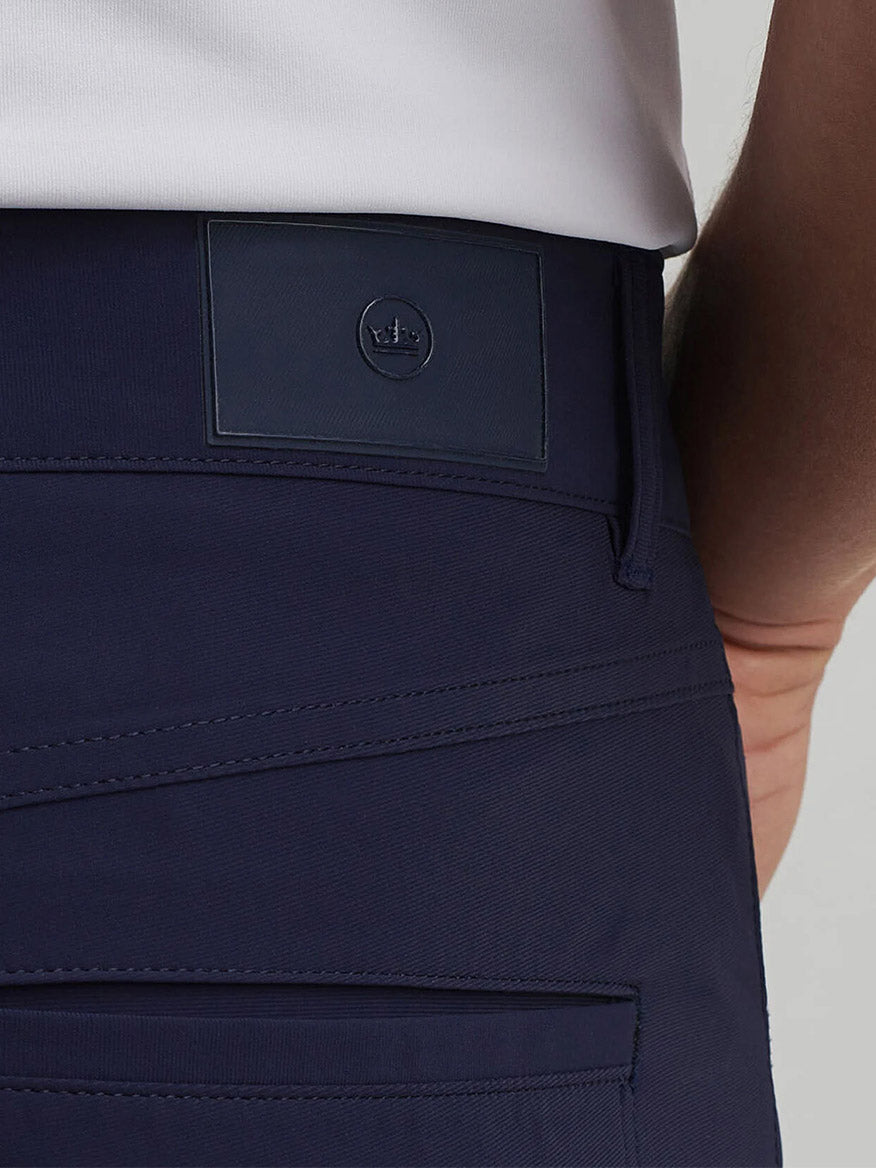 Close-up of a person wearing Peter Millar Bingham Performance Five-Pocket Pant in Navy, made from Italian four-way stretch knit fabric with a rectangular black crown logo patch near the waistband, paired with a white shirt.