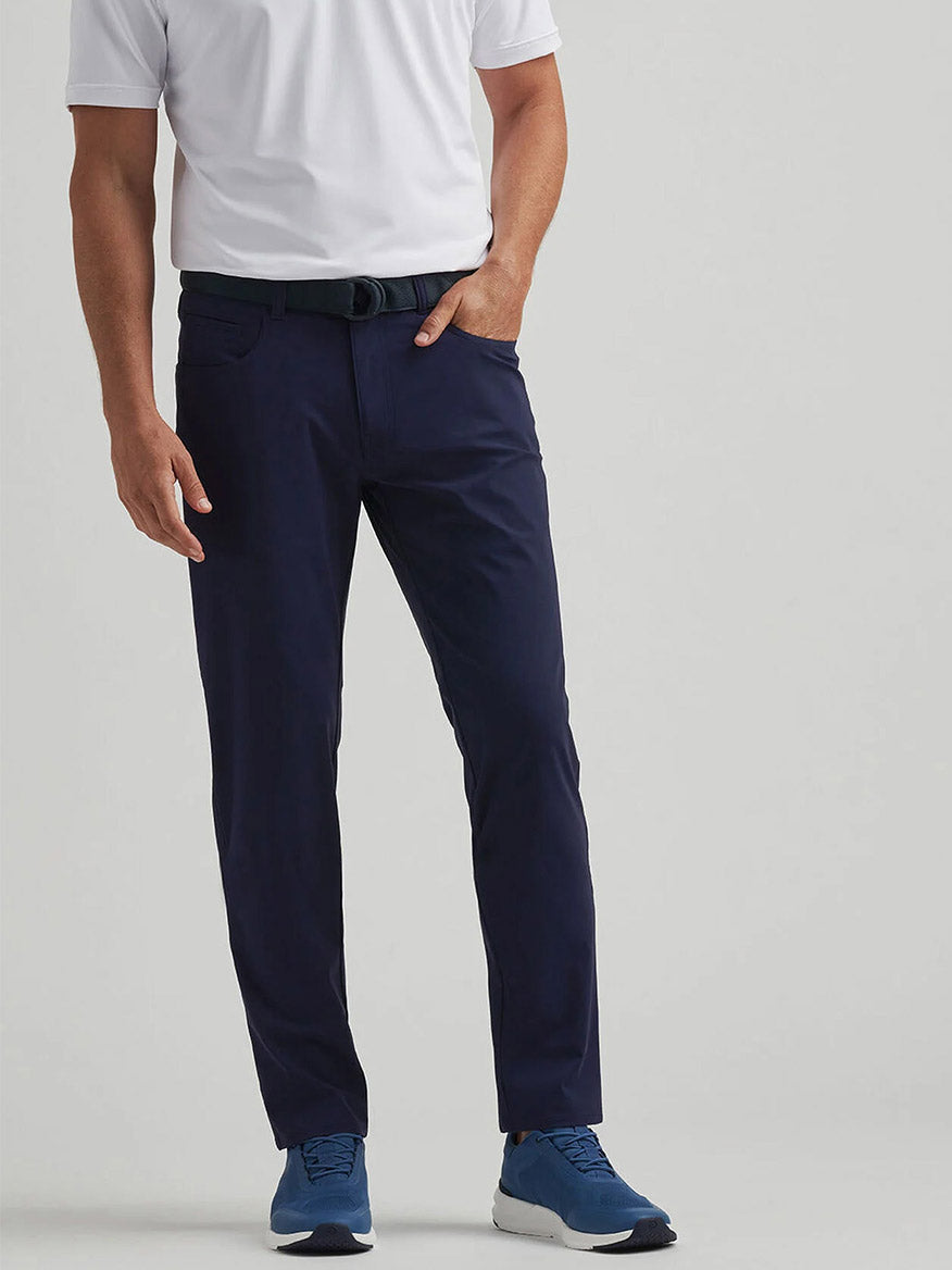 A man stands against a plain background wearing a white polo, the Peter Millar Bingham Performance Five-Pocket Pant in Navy, a black belt, and blue sneakers.