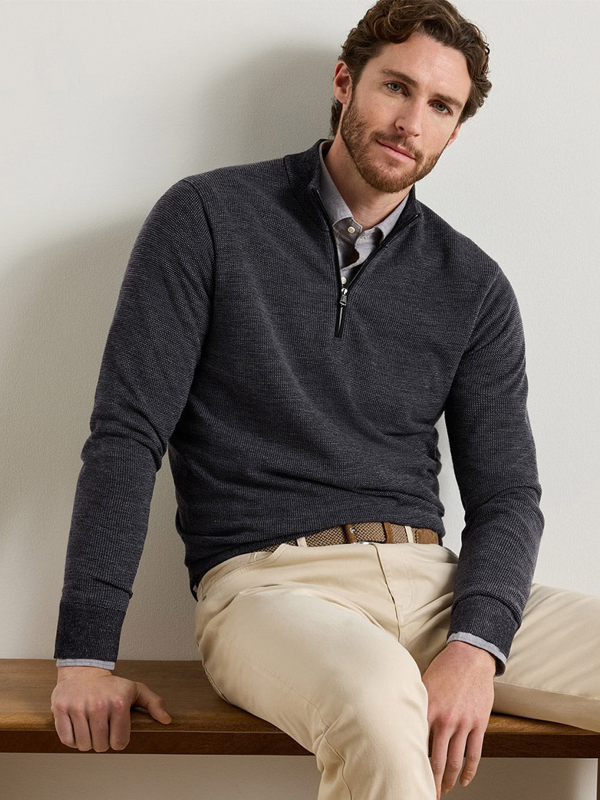 A man wearing a charcoal Peter Millar Blair Birdseye Quarter-Zip Sweater over a light-colored button-down shirt and tan trousers.