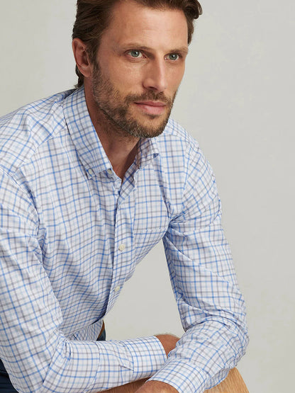 A brown-haired, bearded man wearing a Peter Millar Calgary Crown Lite Cotton-Stretch Sport Shirt in Blue Poppy leans forward with his hands on his knee against a light gray background.