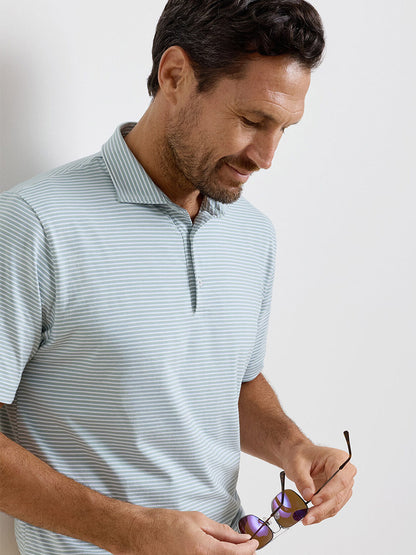 Man wearing a light blue striped Peter Millar Calvert Crown Flex Cotton Polo in Grandview looks down while holding sunglasses against a plain white background.