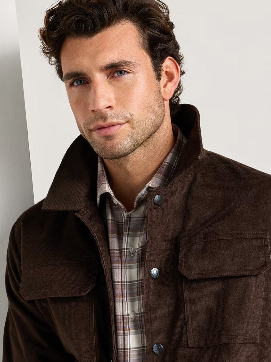 A man with short brown hair wears the Peter Millar Camden Corduroy Jacket in French Press over a plaid shirt, standing indoors against a light-colored wall and showcasing effortless lightweight layering.