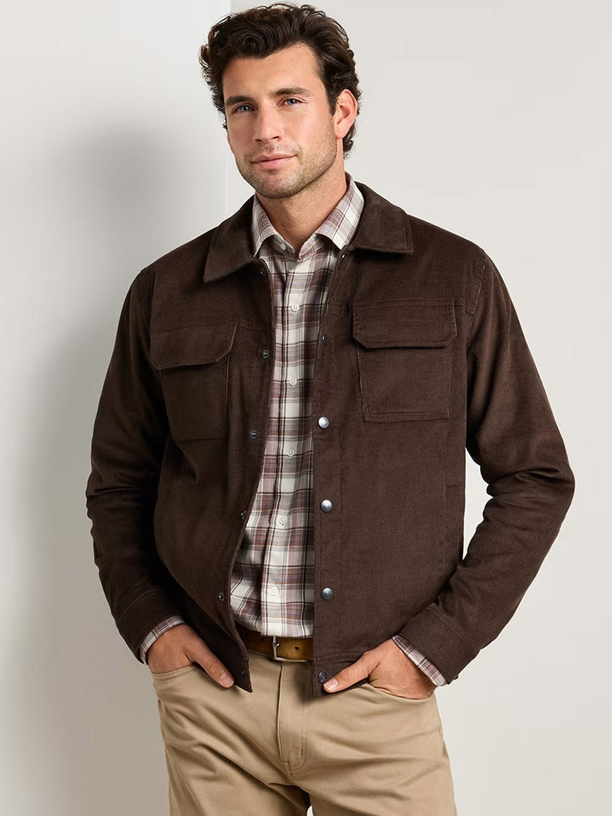 A man stands with hands in his pockets against a plain light background, effortlessly layering the Peter Millar Camden Corduroy Jacket in French Press over a plaid shirt and khaki pants.