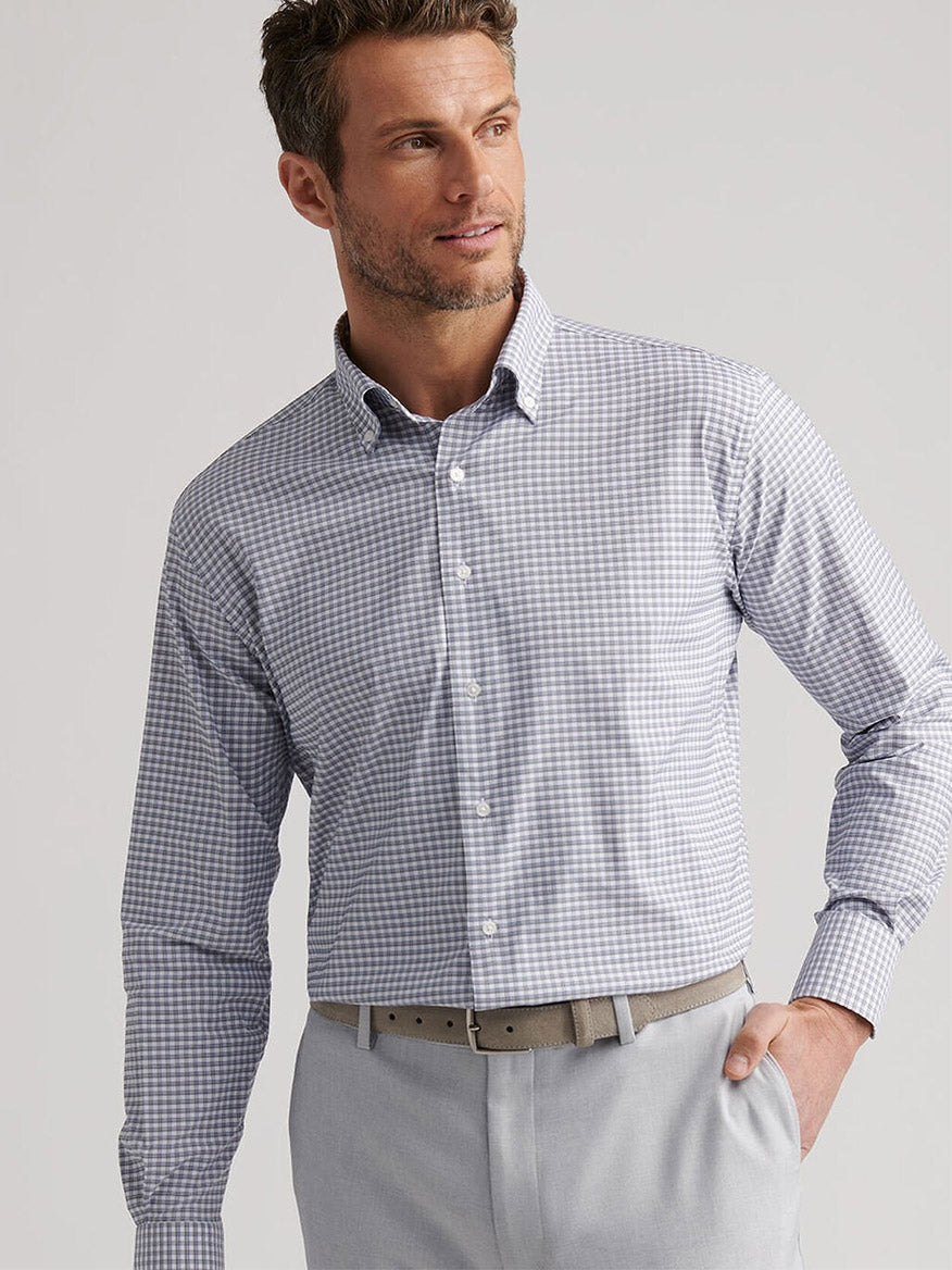 A man wearing the Peter Millar Cashiers Performance Twill Sport Shirt in Oslo Grey, tucked into light gray pants with a beige belt, stands against a plain background.