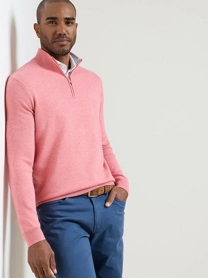 A man in the Peter Millar Coolspun Lite Cotton Quarter-Zip Sweater in Venetian Pink and blue pants leans against a white wall with one hand in his pocket, showing off a classic fit.