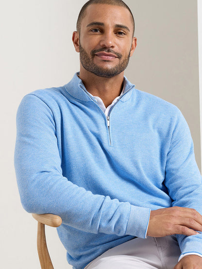 A man with short hair and a beard wears the Peter Millar Coolspun Lite Birdseye Quarter Zip Sweater in Bluebird, featuring moisture-wicking fabric, as he sits on a wooden chair against a light background.