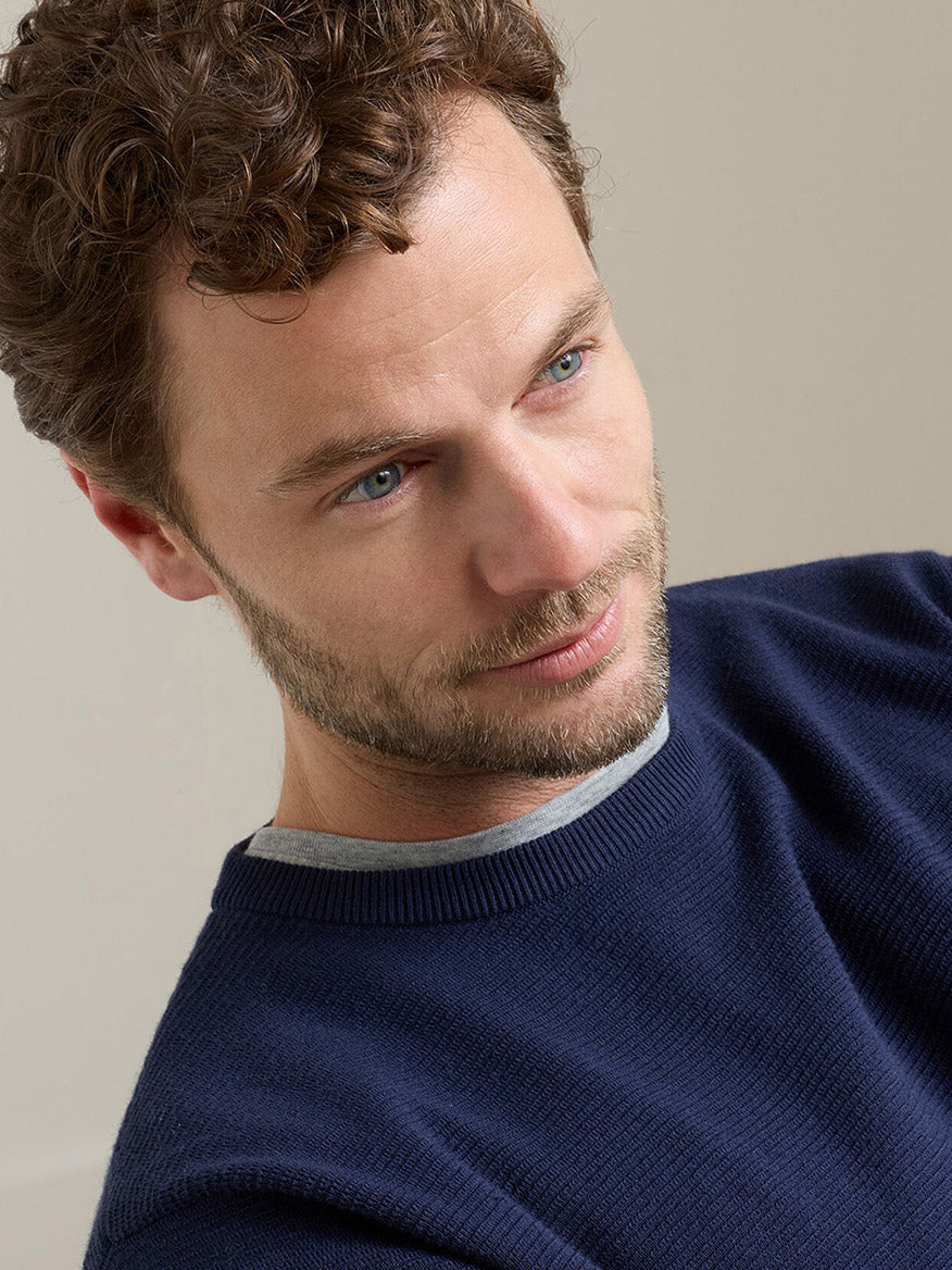 A man with curly brown hair and a beard wears the Peter Millar Coolspun Lite Textured Crewneck Sweater in Navy over a gray shirt, looking slightly to the side against a plain background.