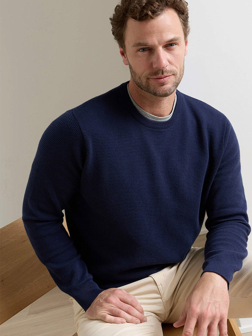 A man with light brown hair sits on a wooden chair, wearing the Peter Millar Coolspun Lite Textured Crewneck Sweater in Navy and light-colored pants, looking at the camera.