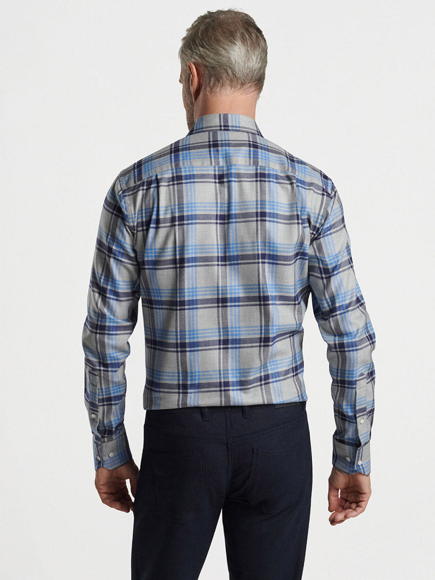 A man with gray hair stands facing away, wearing the Peter Millar Coulter Italian Flannel Sport Shirt in Gale Grey, known for its tailored fit and stylish cotton-blend plaid design in gray and blue, paired with dark pants against a plain background.