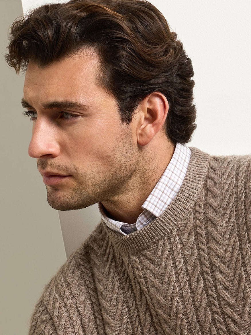 A man wearing a Peter Millar Crescent Cable Crewneck Sweater in Sawgrass over a light-colored checkered button-down shirt.