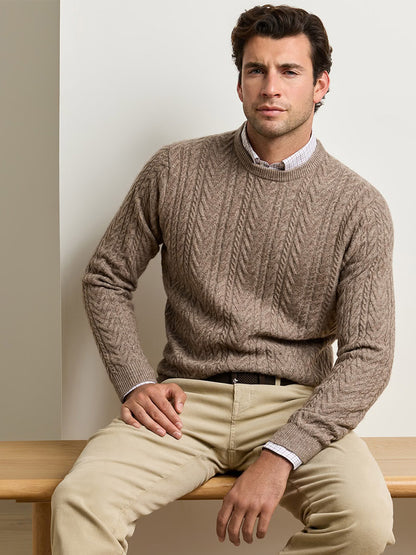 A man wearing a tan Peter Millar Crescent Cable Crewneck Sweater in Sawgrass over a button-down shirt and light-colored trousers.