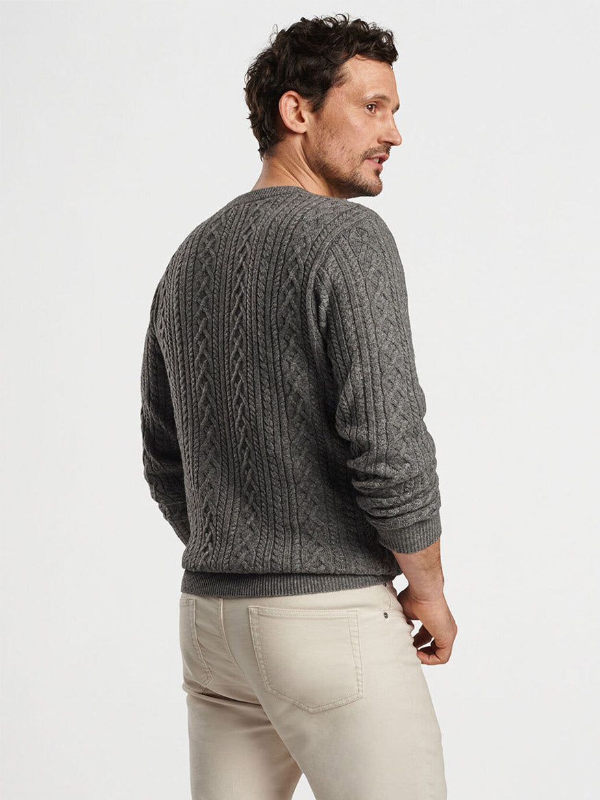 A man wearing the Peter Millar Crescent Ridge Cable Crew in Gale Grey sweater paired with beige pants stands against a plain background, looking to the side.