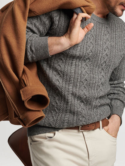 A person wearing the Peter Millar Crescent Ridge Cable Crew in Gale Grey, along with beige pants and a brown belt, holds a brown coat over their shoulder, creating the ideal ensemble for 3-season wear.