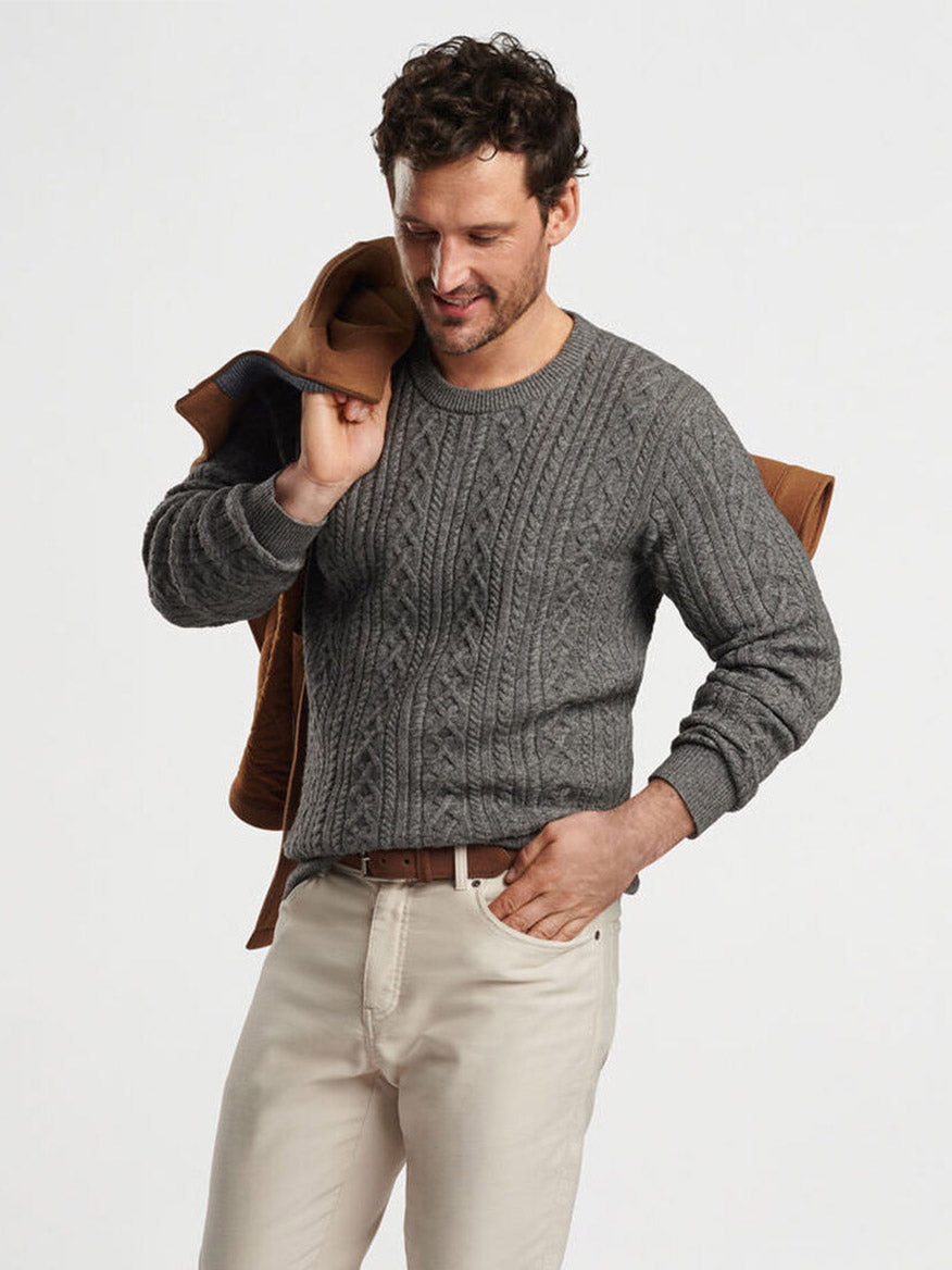 A man in a Peter Millar Crescent Ridge Cable Crew in Gale Grey, crafted from cozy Merino wool, holds a brown jacket over his shoulder, standing against a plain background.