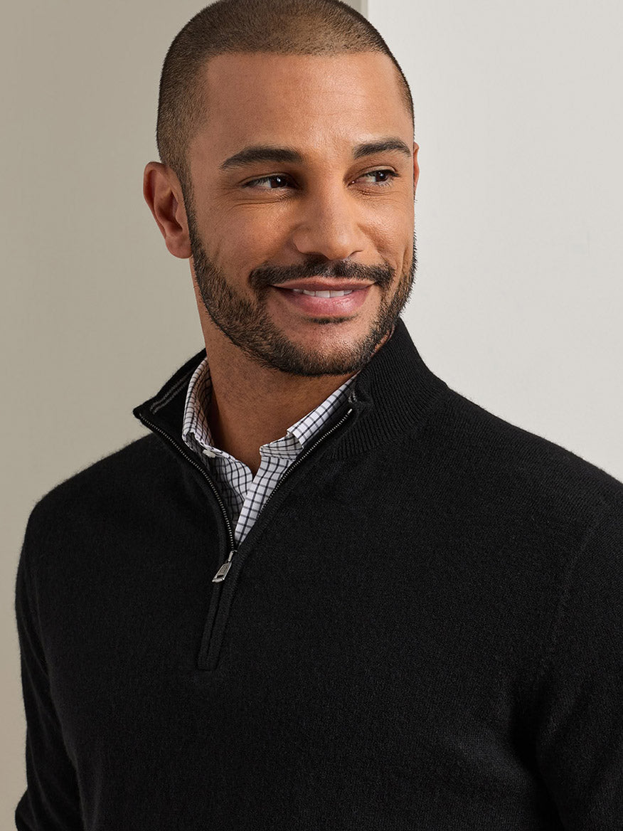 A man wearing a black Peter Millar 25th Anniversary Cashmere Quarter-Zip Sweater over a checkered collared shirt.
