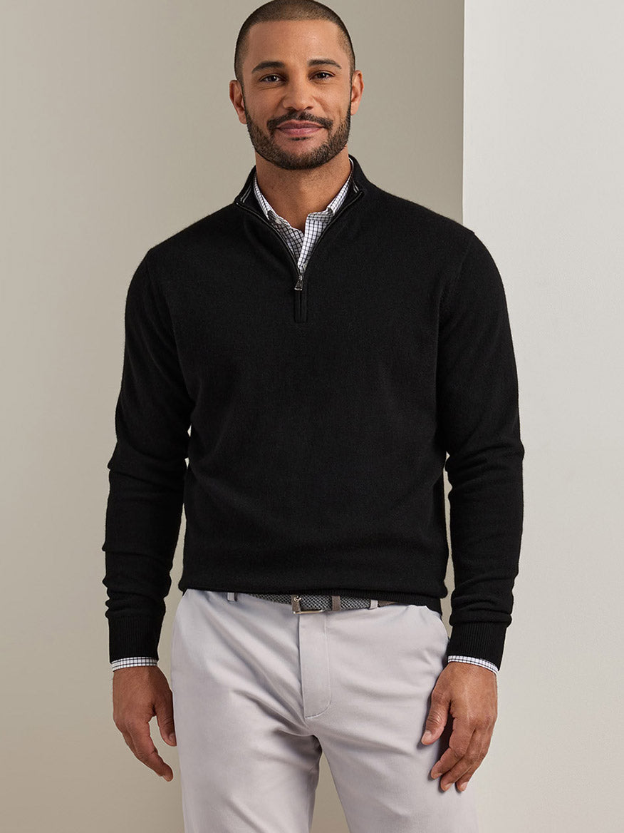 A man wearing a Peter Millar 25th Anniversary Cashmere Quarter-Zip Sweater in Black over a checkered collared shirt and light gray trousers.