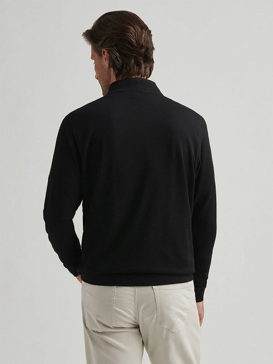 A man stands with his back to the camera, wearing the Peter Millar Crown Comfort Pullover in Black and light-colored pants against a plain background.